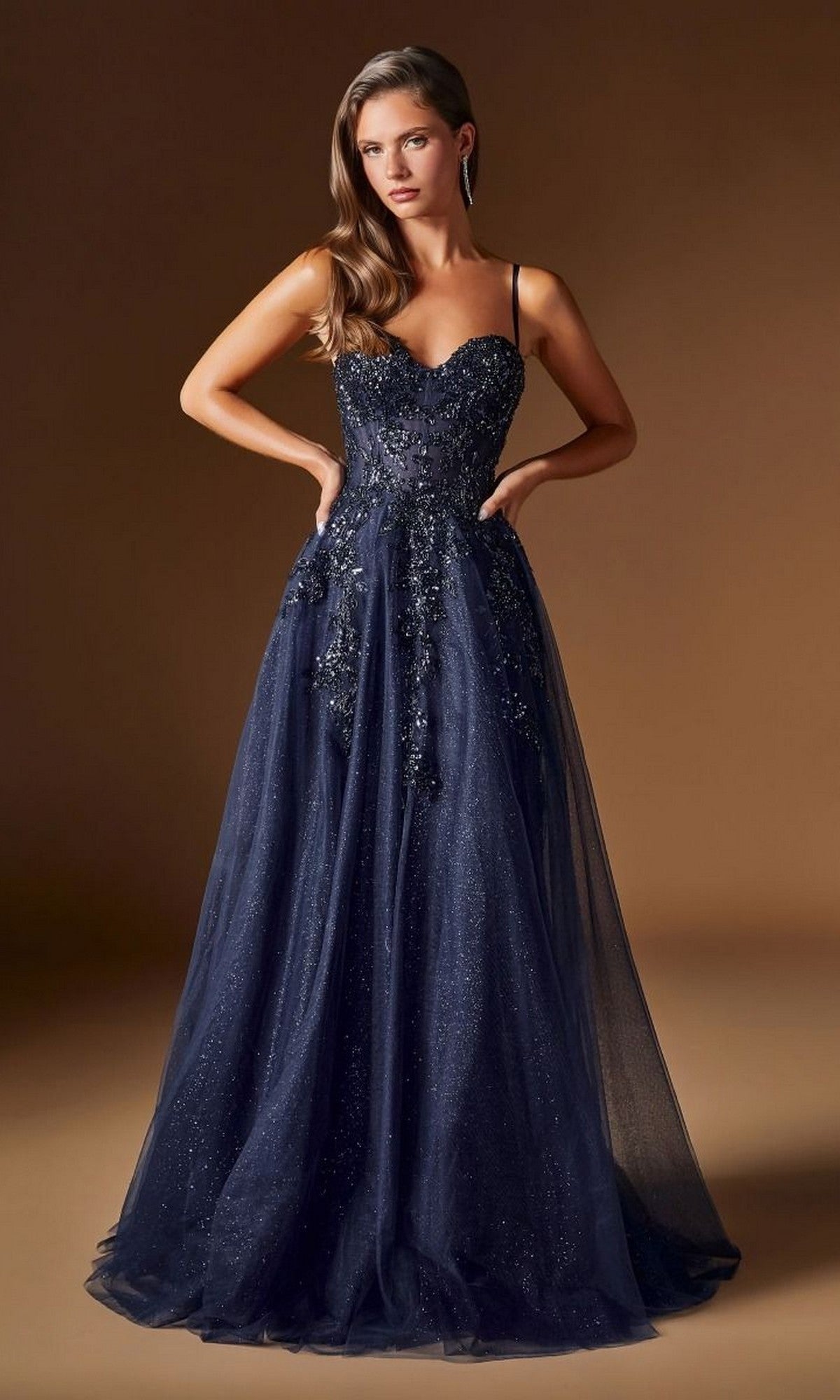 A woman poses with hands on hips in the Ladivine C177 long prom dress, showcasing its navy blue color, thin straps, embellished bodice, and sparkly accents against a plain brown background.